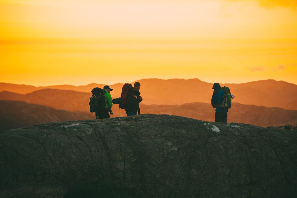3 mountaineers with backpacks hiking on a mountain top at dusk.