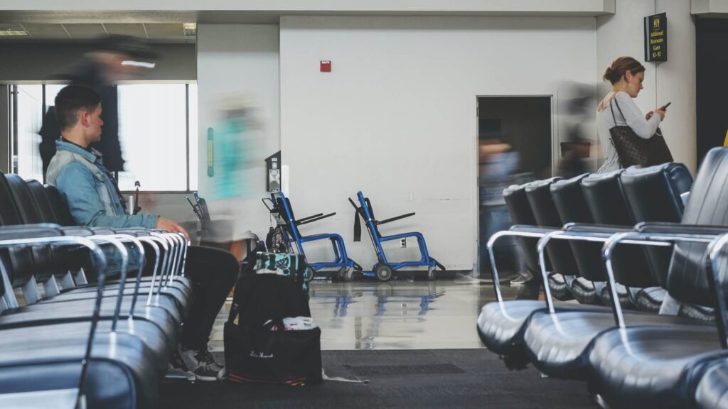 A man in an airport sitting down while waiting for his flight and a woman walking past focusing on her phone