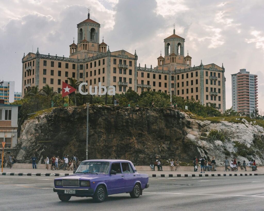 A castle on a hill in Cuba, people walking on the pedestrian lane and a car parked at the intersection of the road