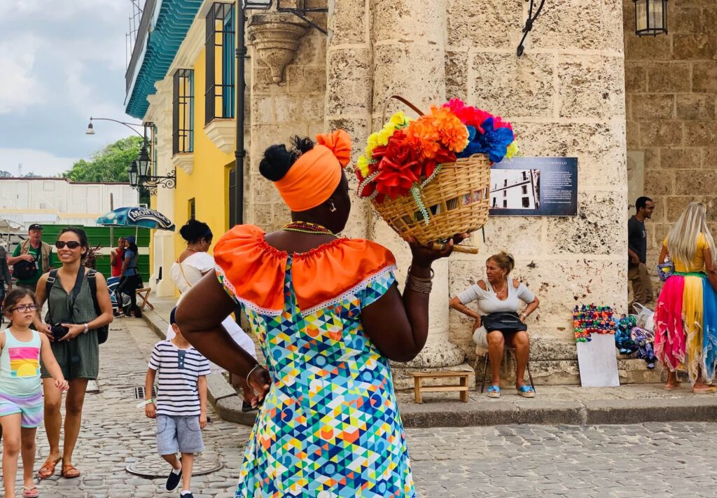 A black woman holding a handcrafted basket containing some flowers as well as other people visiting Cuba